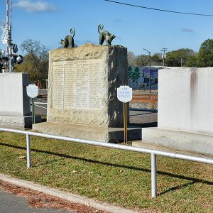 Three engraved stone monuments near train crossing with signs and metal railing around them