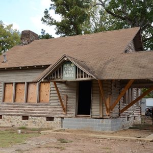 Log cabin building with boarded up windows and car port on one side and horses tied up on the other side