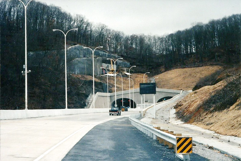 Two highway tunnels through a tree-covered mountainside with street lamps
