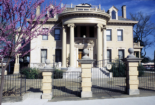 Three-story stone building with entrance covered with round covering and four columns behind black iron fence with three columns and a tree with purple flowers