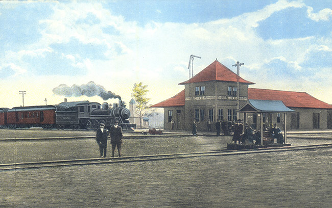 Steam train with cars arriving at multistory depot building with patrons in rail yard