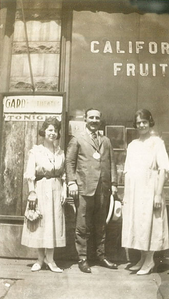 White man in suit and tie with two white women outside a storefront with "California Fruit" written on the window