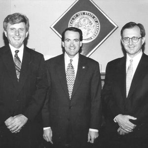 Three white men in suits with state seal and flag behind them