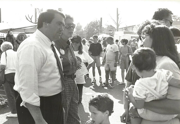 White man in shirt and tie alongside woman in sunglasses crowd talking to white woman holding a child amid an outdoor crowd