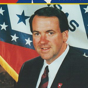 White man in suit and striped tie on "Mike Huckabee Governor" campaign card with Arkansas flag behind him