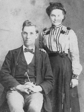 White man in suit and bow tie sitting with white woman in dress standing behind him