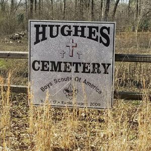 White "Hughes Cemetery" sign on wooden fence in overgrown cemetery