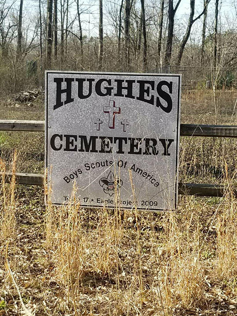 White "Hughes Cemetery" sign on wooden fence in overgrown cemetery