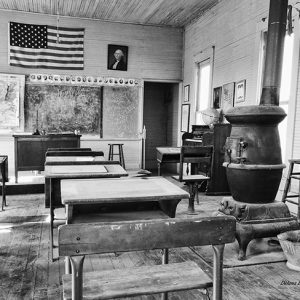 Flag maps and photographs on the walls inside classroom with teacher's desk and rows of student's desks and wood burning stove