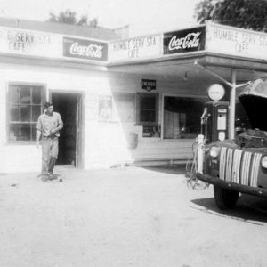 White man standing by service station building as an older white man works on his truck in the foreground