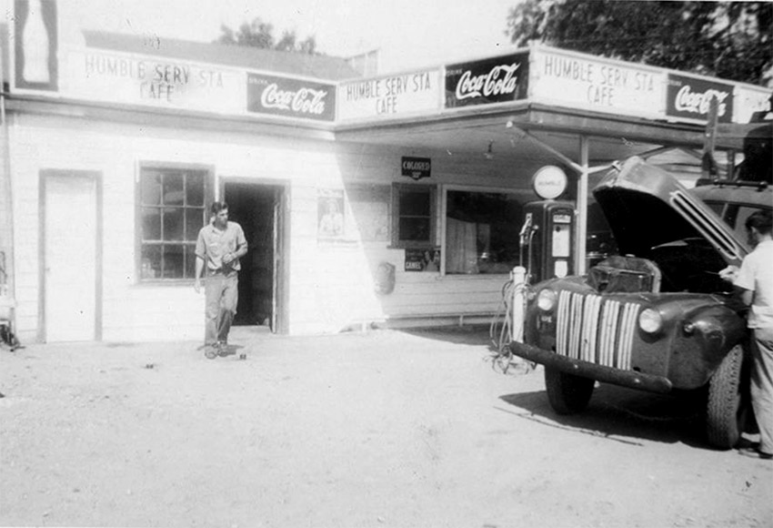 White man standing by service station building as an older white man works on his truck in the foreground