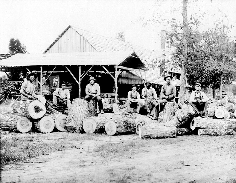 White loggers in hats and overalls at lumber mill with big logs