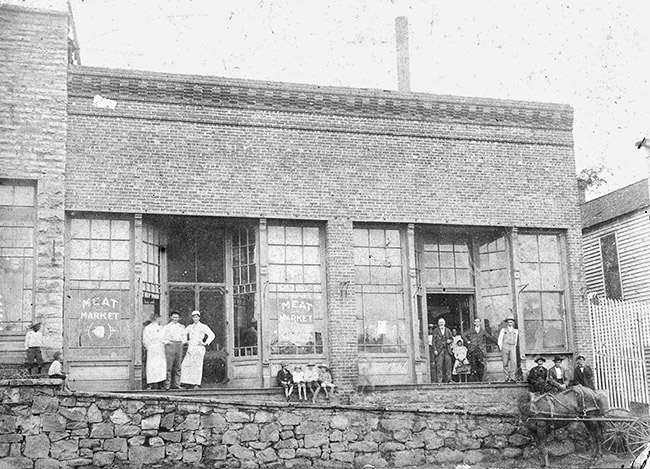 White workers and others standing outside brick building with stone wall and horse drawn wagon