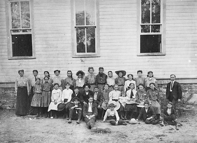White children and teachers posing together outside building with wood siding and rectangular windows