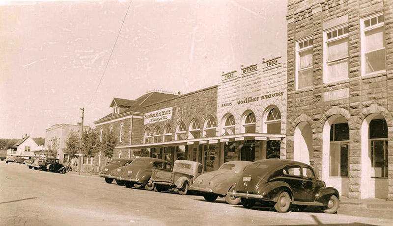 Parked cars sitting outside brick storefront buildings on town street with multistory buildings and house in the background