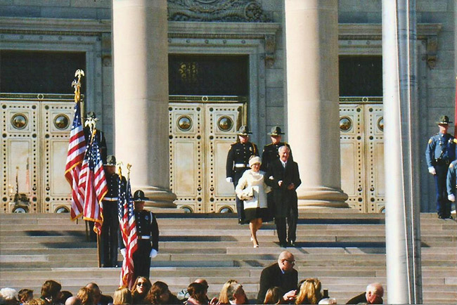 White man in suit and woman in white coat on capitol building steps with color guard policemen and crowd