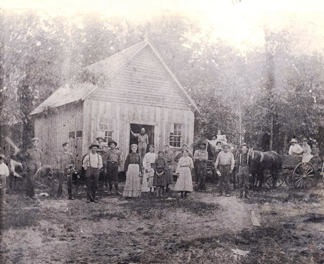 Group of white men women and children standing outside single-story building with A-frame roof and trees in background