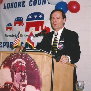 White man in suit and tie speaking at lectern with "Lonoke County Republican Party" banner on wall behind him
