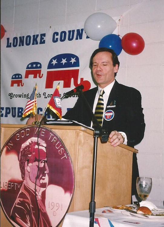 White man in suit and tie speaking at lectern with "Lonoke County Republican Party" banner on wall behind him