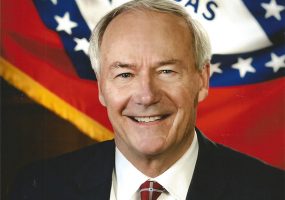 White man in suit and tie smiling with Arkansas flag behind him