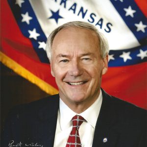 White man in suit and tie smiling with Arkansas flag behind him