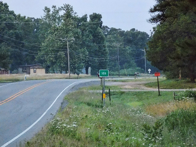 "Ida" road sign on two-lane highway with mobile homes in the background