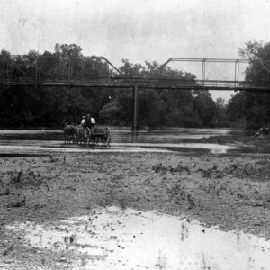 Men crossing a partly dry river bed with horse drawn wagon with steel truss bridge in the background