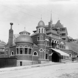 Three-story brick structure with round towers and arched entrance on dirt road with taller building in the background and a horse and buggy in front