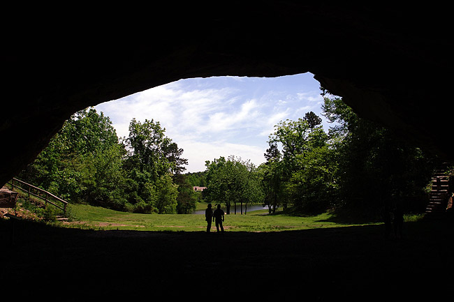 Two people standing at cave entrance with pond and building in the background as seen from inside the cave