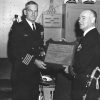 White men in military uniforms with swords holding plaque in room with cabinet behind them