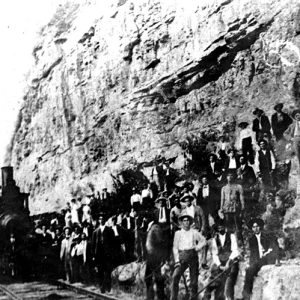 Group of white workers standing on edge of rock wall with steam locomotive on railroad tracks in the foreground