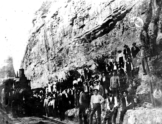Group of white workers standing on edge of rock wall with steam locomotive on railroad tracks in the foreground