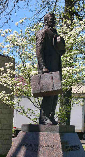 Statue of a man with suitcase in his right hand and hat over his heart on engraved pedestal next to tree with white flowers