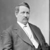 White man with mustache in suit sitting in chair