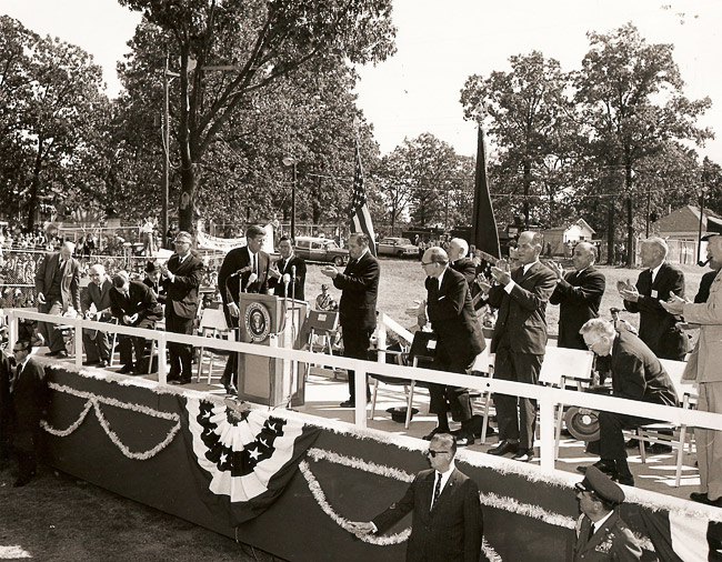 Decorated outdoor stage with white men and women clapping as a white man in suit approaches the lectern