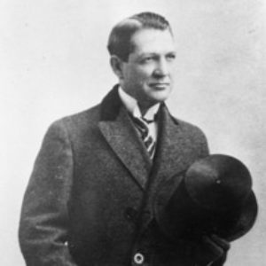 White man with slicked back hair in suit and tie and top coat holding cane and hat