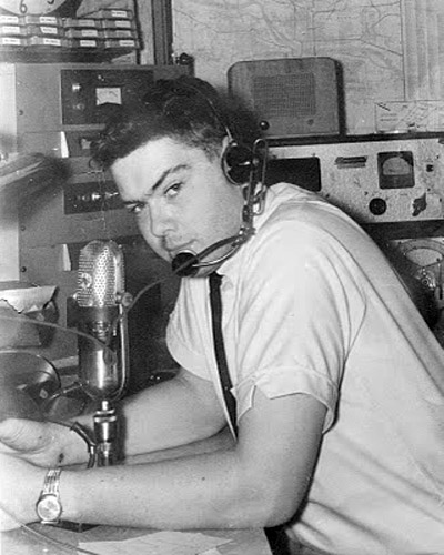 Young white man with headphones in radio booth with recording equipment around him