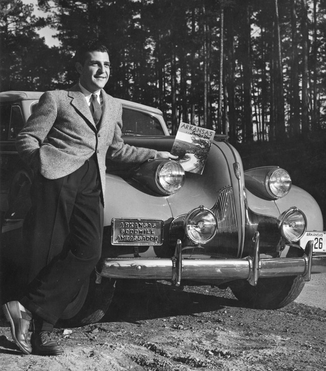 smiling dark-haired white man in tweed jacket dark pants and tie holding a book and standing in front of a shiny car