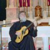 White man with beard in robes playing classical guitar in church with altar behind him