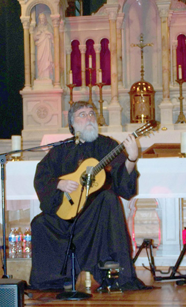 White man with beard in robes playing classical guitar in church with altar behind him
