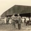 Two white men shaking hands outside airplane hanger with crowd behind them