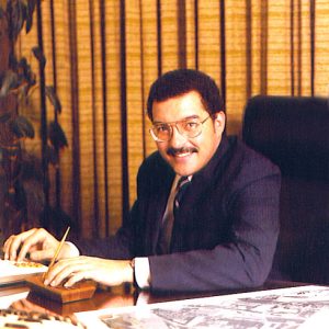 Portrait black man smiling mustache glasses suit and tie at office desk near curtains, plant