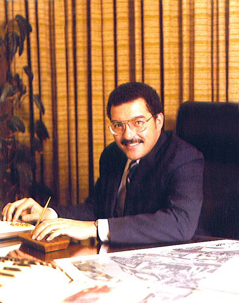 Portrait black man smiling mustache glasses suit and tie at office desk near curtains, plant