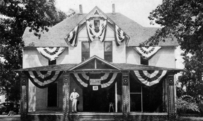 White man posing on front porch of multistory wood frame home that is covered in patriotic banners and a sign saying "Welcome Joe"