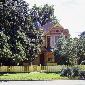 Two-story house painted many bright colors with tower and yellow picket fence with trees