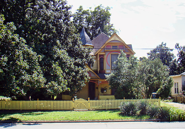 Two-story house painted many bright colors with tower and yellow picket fence with trees