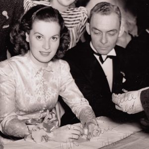 White woman in long sleeved top sitting and smiling at table next to white man in tuxedo