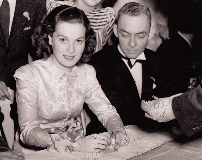 White woman in long sleeved top sitting and smiling at table next to white man in tuxedo