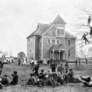 multistory building with covered porch with white students playing in the yard and some in a tree