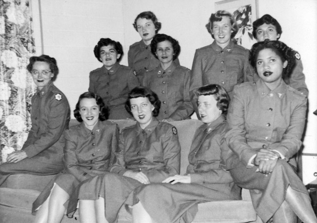 Group photo ten women (nine white, one black) in matching uniforms jackets skirts pose on and around couch indoors